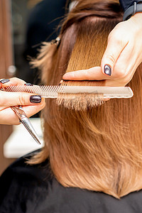 红色发束摄影照片_美发师的手将发束夹在手指之间，在美发沙龙里用梳子和剪刀为年轻女子的长发进行理发，特写。