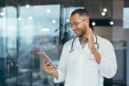 穿着医用白大褂和听诊器的快乐的西班牙裔医生使用平板电脑为患者进行视频通话和在线咨询，在诊所办公楼内工作的男子