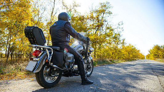 摩托车背景摄影照片_摩托车司机在秋季道路上骑着定制直升机自行车。