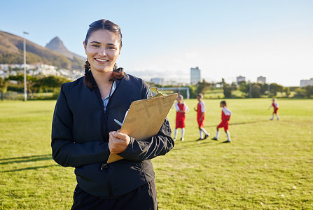 在阳光明媚的日子里，足球教练、初级运动和带剪贴板的妇女在足球场或户外球场指导孩子们。