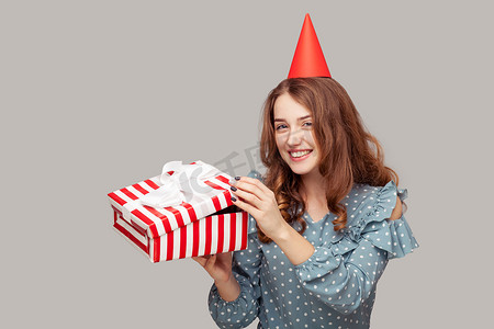 派对帽摄影照片_戴着滑稽派对帽的积极漂亮女孩打开礼盒，看着相机，微笑着对礼物感到满意，享受生日