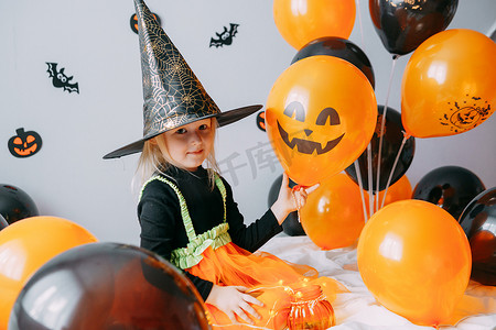 书香飘万家摄影照片_儿童万圣节-一个戴着女巫帽的女孩和一个狂欢节服装，家里有通风的橙色和黑色气球。
