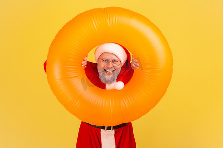 身穿圣诞老人服装、留着灰胡子、微笑的老人手里拿着橙色橡胶圈，表情愉快地看着相机，冬季之旅。