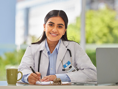 一位戴着听诊器的年轻印度医生的画像坐在办公室里，她坐在办公桌前开处方。