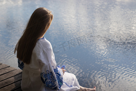 一个穿着乌克兰刺绣衬衫的女孩坐在码头上，湖水中倒映着云彩。