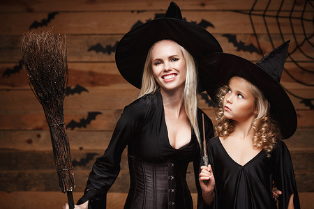 万圣节概念-特写美丽的白种人母亲和她的女儿在庆祝万圣节的女巫服装中与弯曲的南瓜在木制工作室背景上的蝙蝠和蜘蛛网上合影。