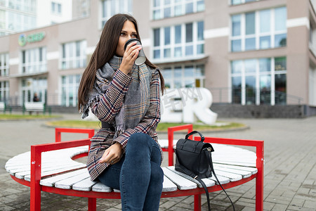 一位身穿秋装的年轻女子坐在长椅上，在建筑物的背景下喝咖啡