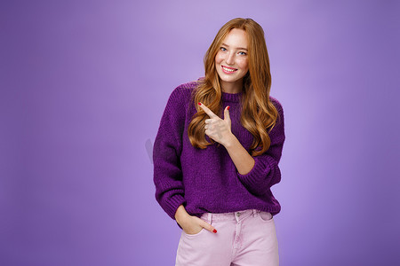 迷人而时尚的女性女性，长着天然红头发，身穿紫色毛衣，手放在口袋里，指着左上角，展示她在紫色墙上做发型或化妆的地方