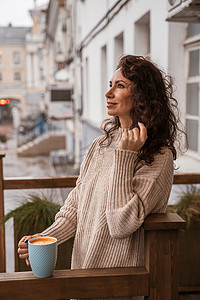 一位身穿米色毛衣、手里拿着蓝色杯子的中年妇女在阳台上的一家街边咖啡馆里