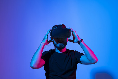 留着胡子的年轻男子戴着 VR 眼镜玩电子游戏，站在蓝色渐变背景的霓虹灯下体验虚拟现实。