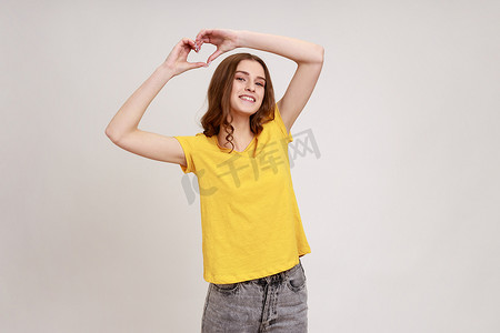 快乐美丽的少女，棕色波浪发，穿着 T 恤，举起双手，展示心形手势，调情，表达爱意。