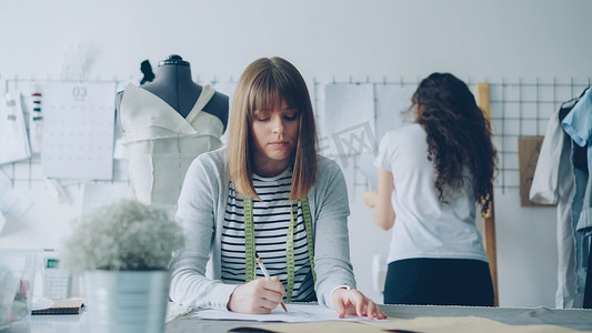 年轻的服装设计师正忙着用铅笔画草图，而她的同事正在设计工作室里观看钉在墙上的图片。