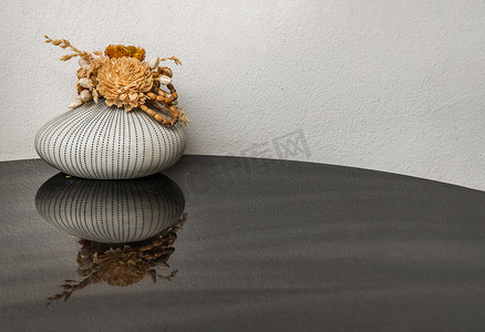 陶瓷业摄影照片_在闪亮的黑色桌面上反射的小手工陶瓷花瓶中的干花。