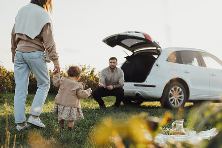 Suv摄影照片_年轻的家庭享受公路旅行，母亲和父亲带着小女儿在户外野餐，背景是 SUV 车