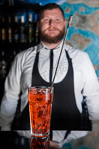 酒吧柜台一位留着胡子的酒保面前，玻璃杯中的红色酒精饮料鸡尾酒和酒吧勺子