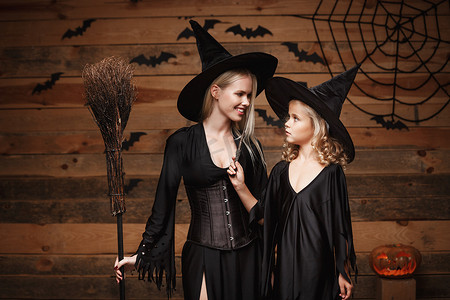 万圣节概念-欢快的母亲和她的女儿穿着女巫服装庆祝万圣节，在木制工作室背景下与蝙蝠和蜘蛛网上的弯曲南瓜合影。
