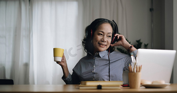 听歌养生摄影照片_戴着耳机的快乐亚洲老年女性在舒适的家客厅里用笔记本电脑听歌