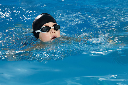 泳帽摄影照片_学龄前男孩戴着泳帽和护目镜学习如何在游泳池里游泳