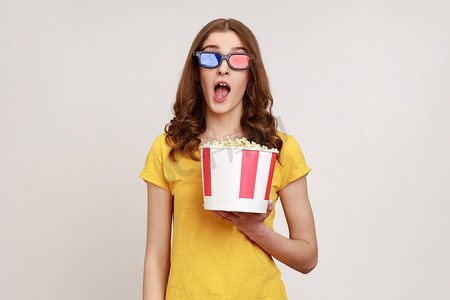 惊讶的年轻女性戴着 3D imax 眼镜观看电影，拿着爆米花，观看精彩的电影，张开嘴，穿着黄色 T 恤。