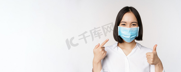 戴着医用面具的亚洲女孩的画像竖起大拇指，指着她的 covid 保护，站在白色背景上