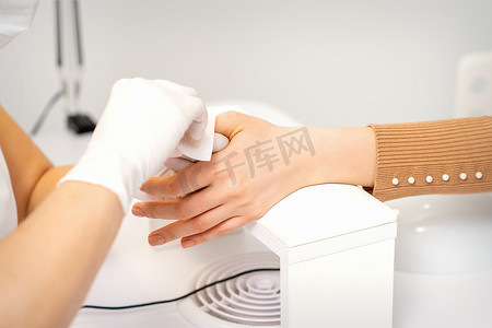 沙龙里一位戴着白色防护手套的美甲师的手用餐巾纸擦拭女性指甲。