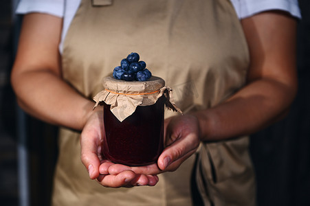 一罐蓝莓果酱，在工艺纸盖上放着新鲜成熟的浆果，在一位妇女、一位糖果家庭主妇手中