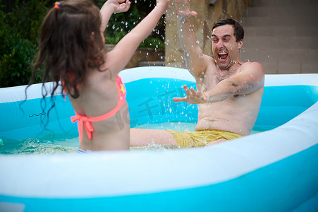 在温暖阳光明媚的夏日，慈爱的父亲和女儿一起泼水，在充气泳池里玩耍。