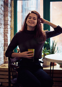 美丽、严肃、时尚、聪明的女孩坐在咖啡馆的窗边，微笑着，喝着健康的纯素冰沙或拿铁。