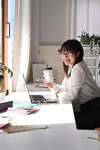 戴着眼镜、开朗的亚洲女员工坐在办公桌前的笔记本电脑前，对着镜头微笑