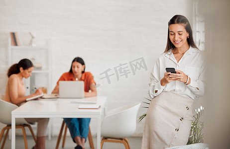 一位快乐的年轻西班牙裔女商人在办公室里用手机发短信，背景是她的同事。