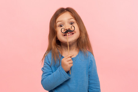 小女孩拿着纸胡子，对着镜头微笑，无忧无虑的孩子玩耍的肖像