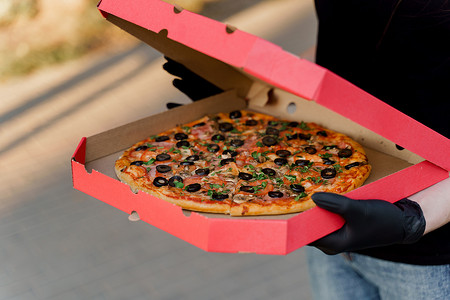 戴着黑色医用手套的食品快递员拿着装有比萨饼店披萨的红盒子。