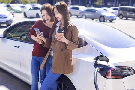 快乐的年轻女子站在电动汽车附近的城市停车场，从小城市车站给汽车电池充电，观看智能手机并检查信息