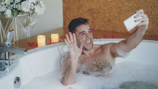 年轻英俊的男子流行博主正在使用智能手机在日间水疗中心的热水浴缸中录制视频。