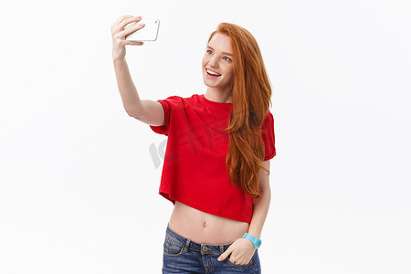形象墙照片墙摄影照片_白墙背景中孤立的年轻快乐红发女性的形象使自拍看起来像相机