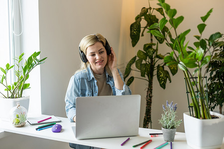 照片中，迷人的积极人士坐在桌面后面，看起来使用笔记本电脑，在室内工作心情很好