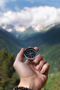 罗盘二十四山摄影照片_一只手拿着罗盘，背景是史诗般的雪山，山上有云，脚下有森林，特写