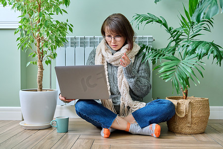 穿着暖和衣服的中年女性靠近暖气片，使用笔记本电脑进行视频通话