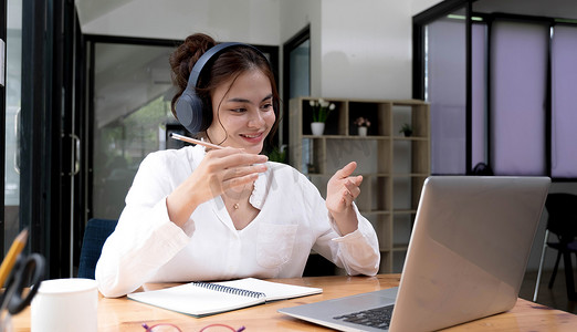 戴耳机的亚洲年轻女性微笑着迎接在笔记本电脑上的网络摄像头虚拟对话中交谈，戴无线耳机的快乐女性在电脑上进行视频通话，在线咨询客户