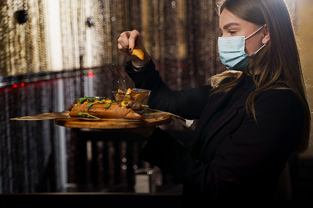烤热狗摄影照片_戴着医用面具的侍者，在木质背景上放着烤热狗，配黄芥末和番茄酱。