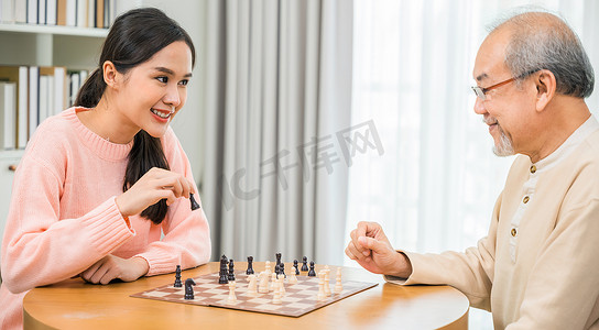 美丽的年轻微笑女人坐在家里与年长的老人下棋，玩得很开心