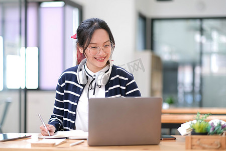 交谈交谈摄影照片_年轻成年快乐微笑的西班牙裔亚裔学生戴着耳机在大学校园或虚拟办公室使用笔记本电脑在在线聊天会议上交谈。