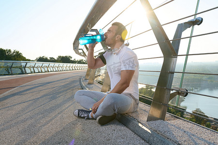 一位肌肉运动员在玻璃城市桥上剧烈运动后喝水和放松的侧面肖像。
