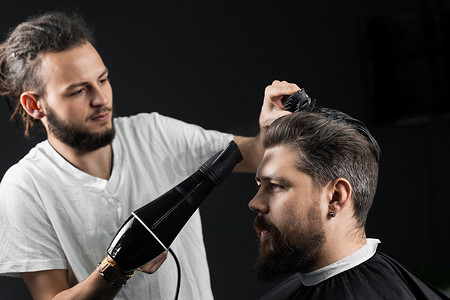 男人吹头发摄影照片_理发师在时尚发型后吹干一位英俊的胡须男子的头发。