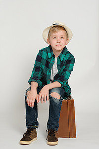 复古邮戳免抠摄影照片_7 岁可爱时尚金发男孩的肖像，戴着帽子、格子衬衫和牛仔裤，坐在复古手提箱上。