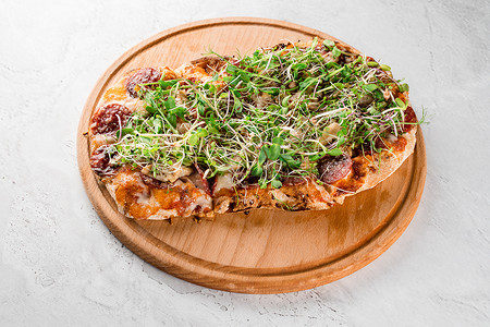 Pinsa romana 配意大利腊肠、奶酪、蘑菇，在白色背景的木板上装饰着微绿色植物。