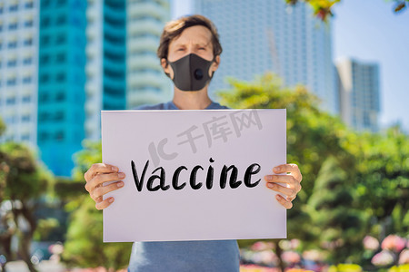 戴医用面具的人可以预防冠状病毒病，他拿着一张海报疫苗手写文字-用白色隔开的字母。