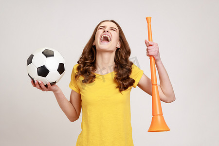 最强少女摄影照片_穿着黄色 T 恤的兴奋的少女拿着喇叭和黑白足球，庆祝最喜欢的足球队在冠军赛上的胜利。