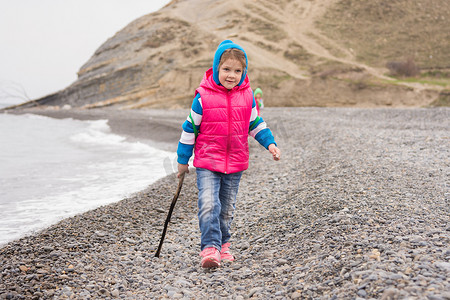 五岁女孩穿着温暖明亮的衣服，手里拿着棍子，在圆石滩上行走