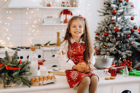 圣诞桌面摄影照片_圣诞厨房里一个快乐的小女孩坐在桌子上，手里拿着饼干